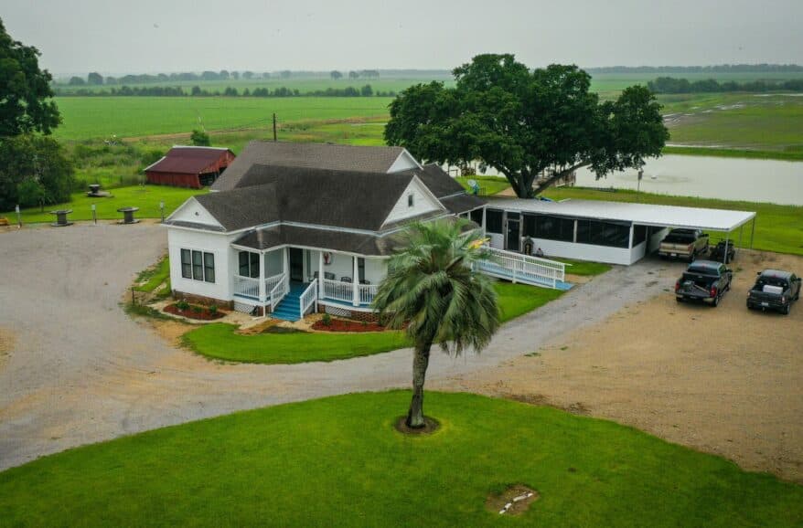 Aerial view of a white house with a porch, surrounded by green fields and parked cars.