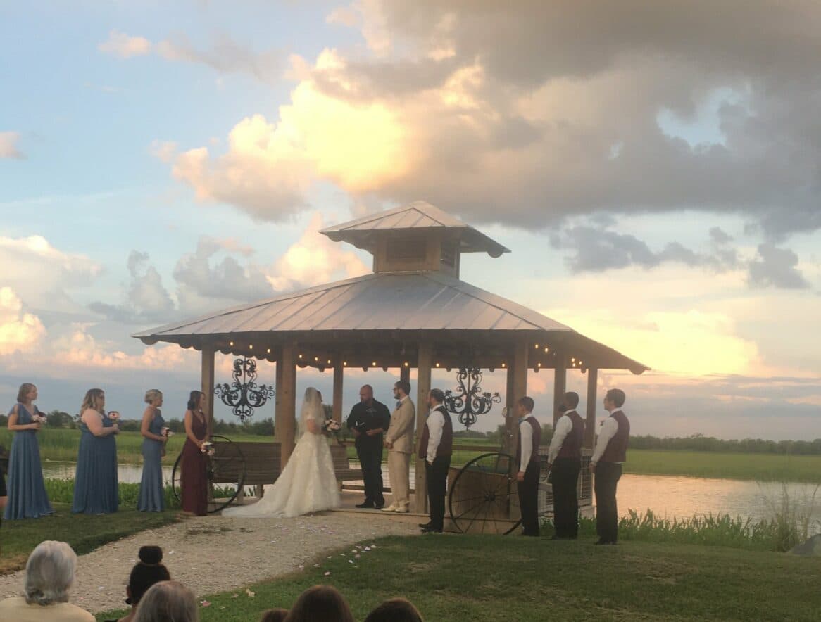 A bride and groom exchange vows under a gazebo surrounded by wedding guests and a scenic sunset.