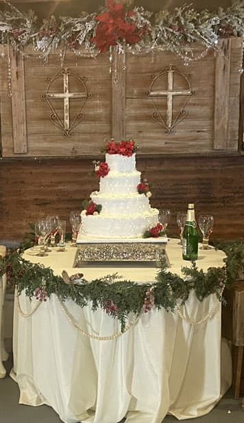 A decorated wedding cake with red roses sits on an elegantly draped table surrounded by glasses and festive greenery.