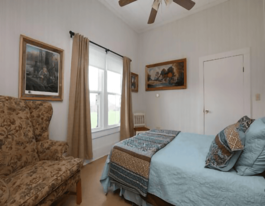 A cozy bedroom with a bed, decorative pillows, armchair, and framed artwork on the walls.