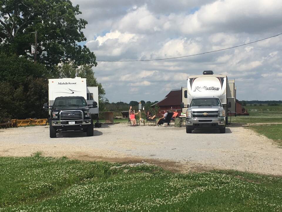 Two RVs parked on a gravel area with people sitting nearby in a rural setting.
