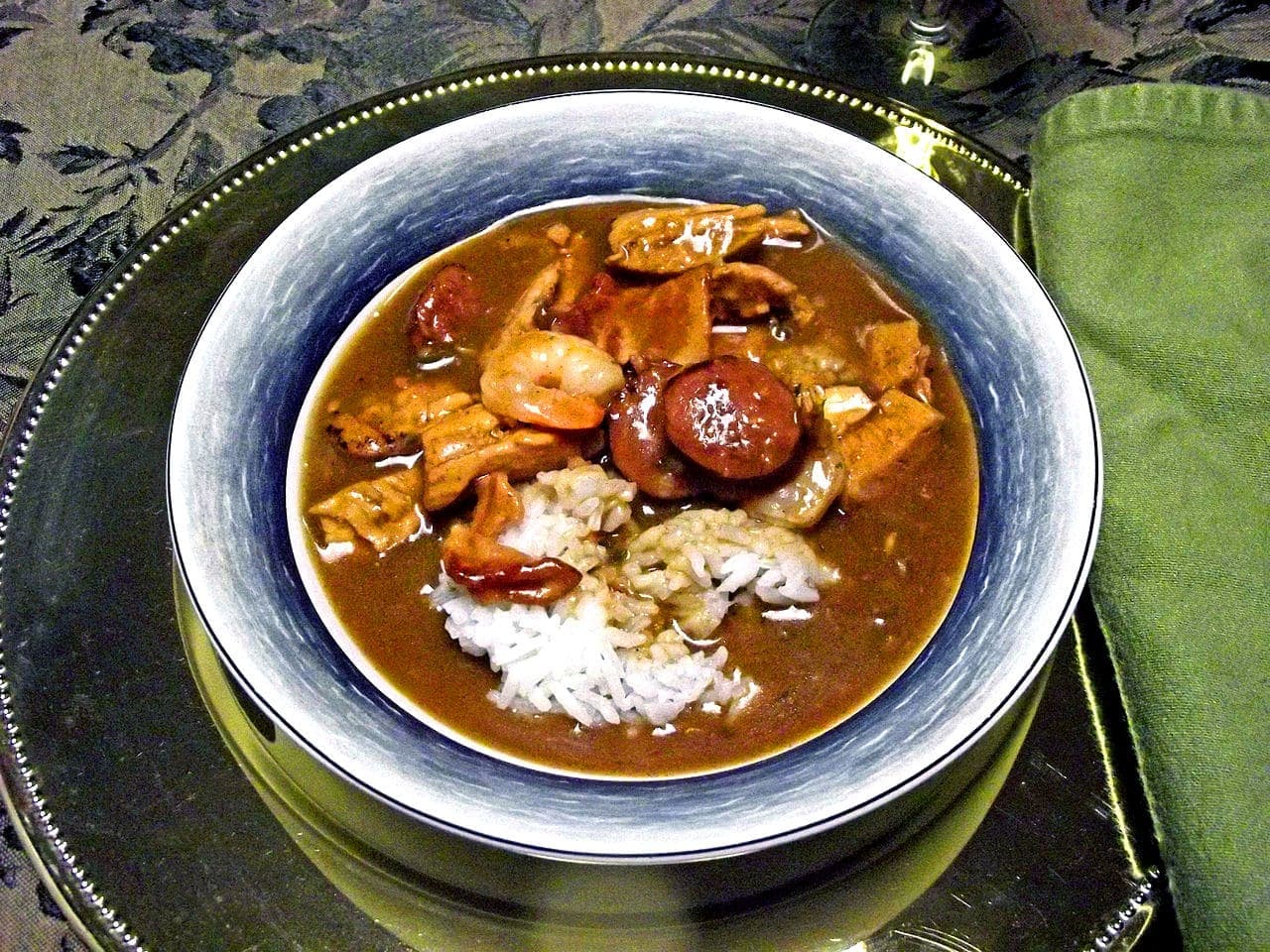 A bowl of gumbo with shrimp, chicken, sausage, and rice served on an elegant table setting.