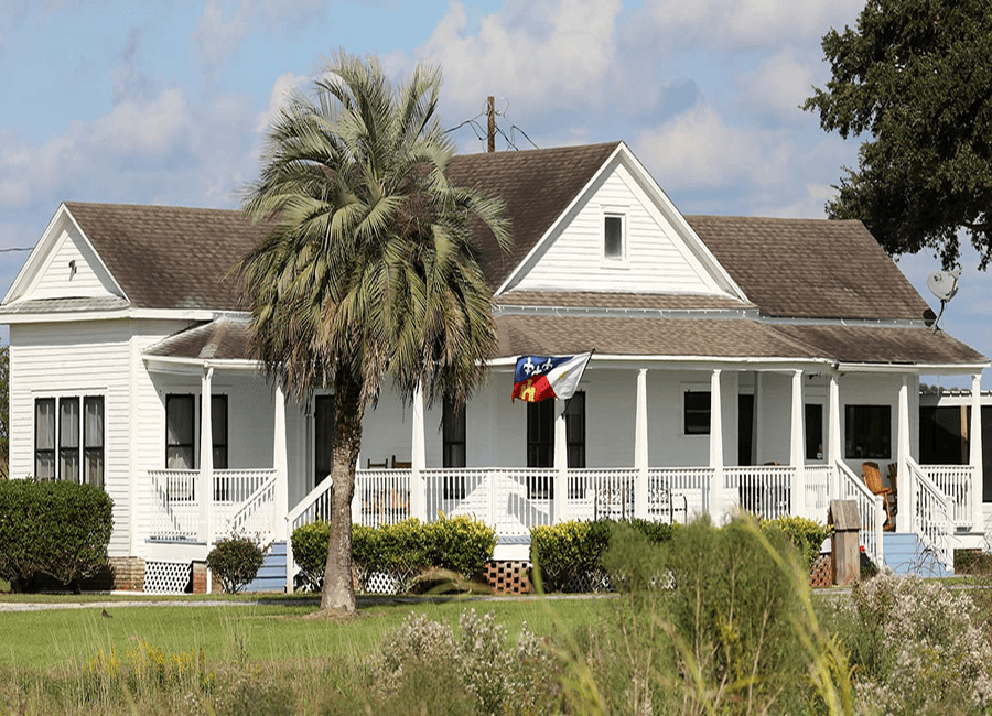 A white house with a porch and Texas flag, surrounded by palm trees and greenery.