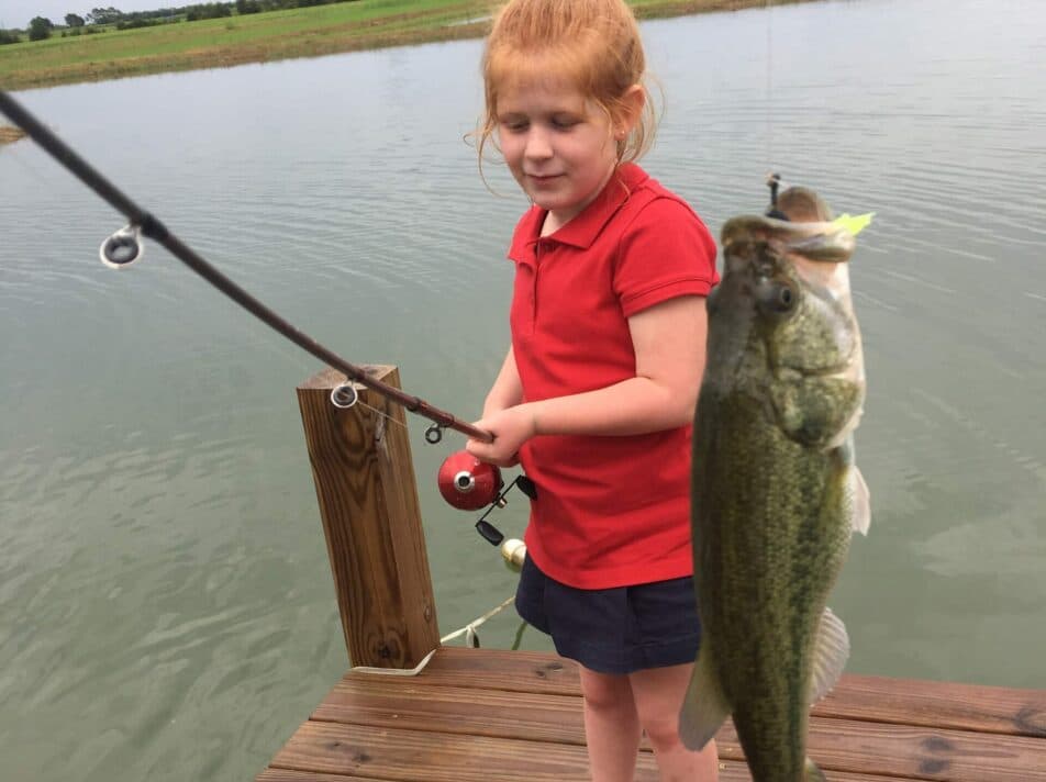 A young girl in a red shirt proudly holds a fishing rod with a caught fish while standing on a wooden dock by a lake.