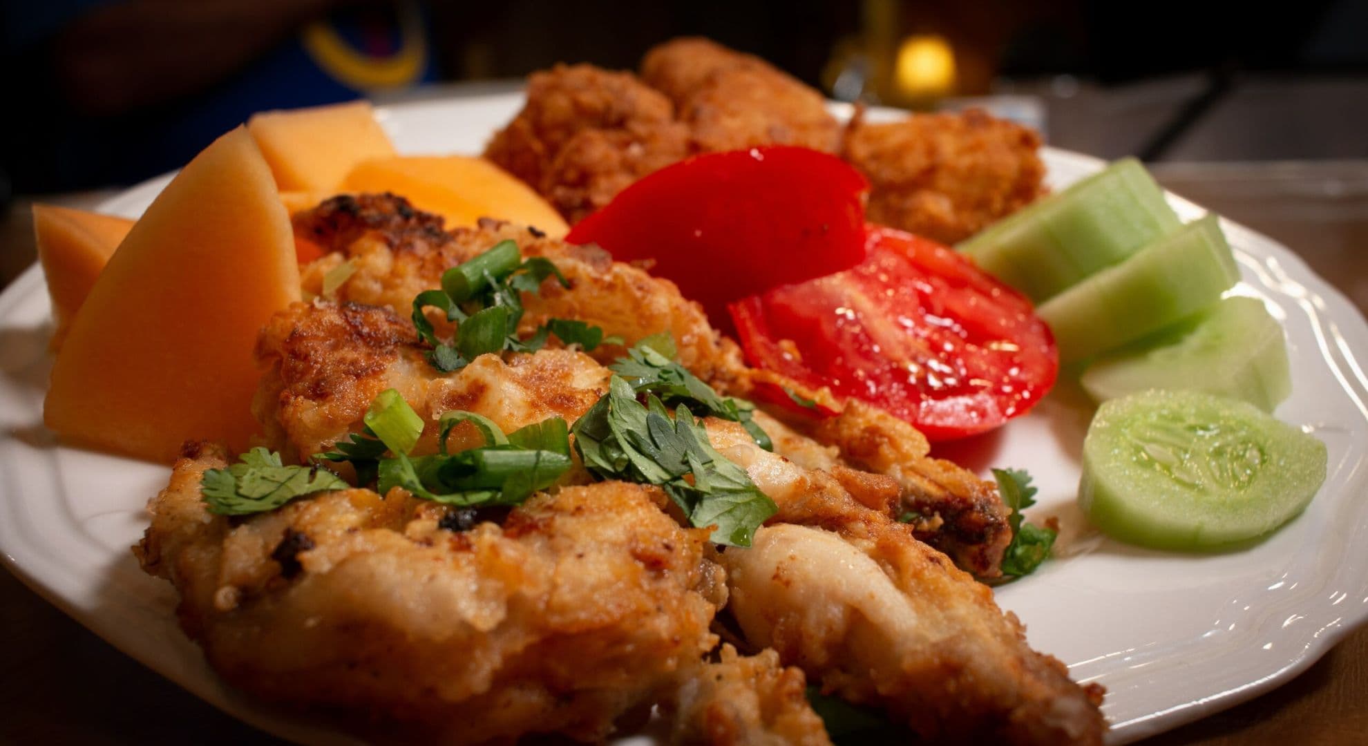 A plate of fried chicken drumsticks garnished with cilantro, accompanied by slices of cucumber, tomato, and cantaloupe.