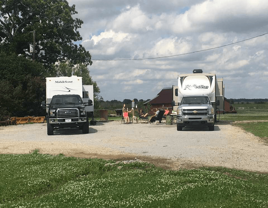 Two pickup trucks parked in front of RVs with people sitting in chairs nearby on a gravel lot.