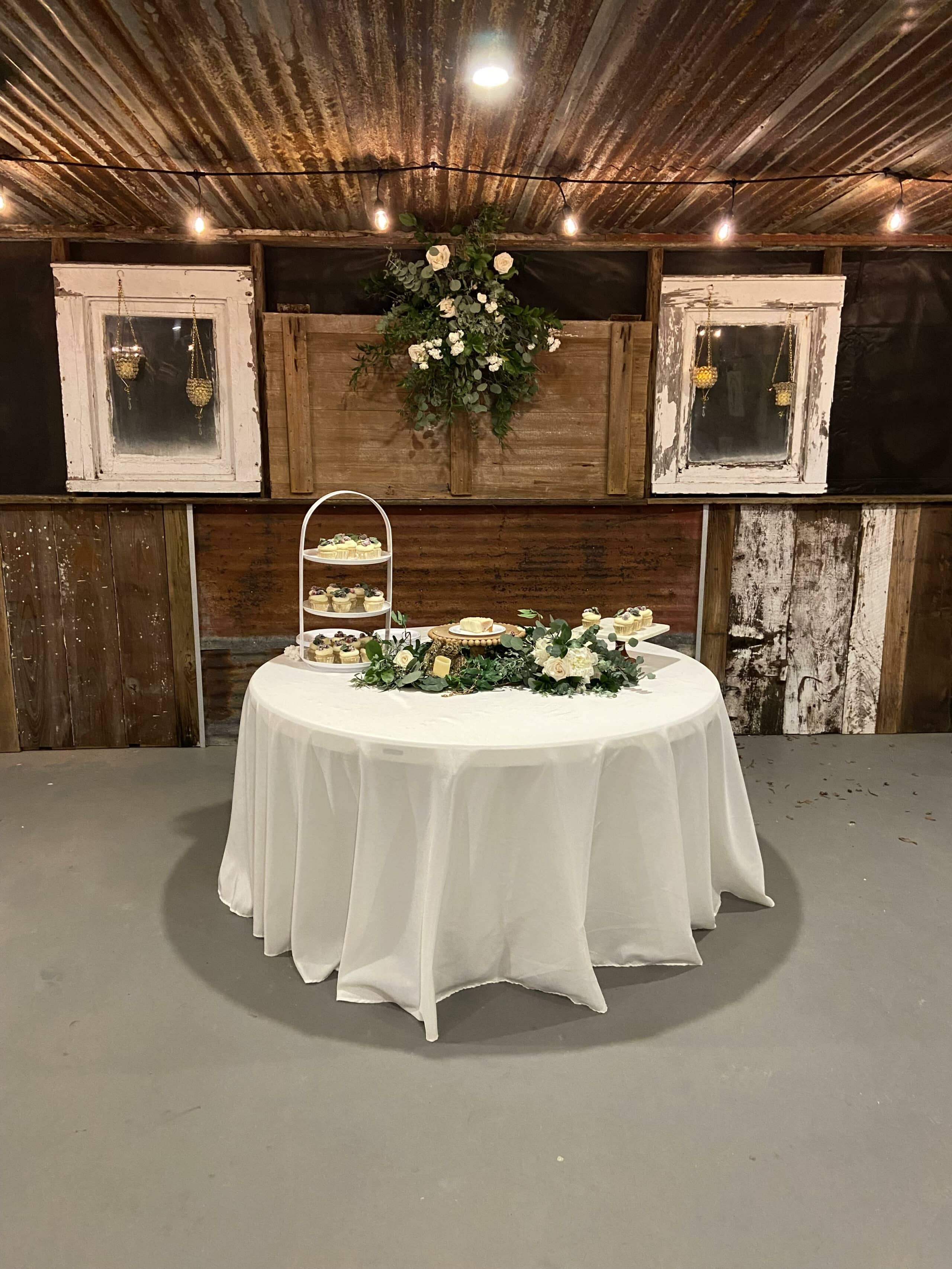 A decorated table with a white cloth features cakes and floral arrangements against a rustic wooden backdrop.