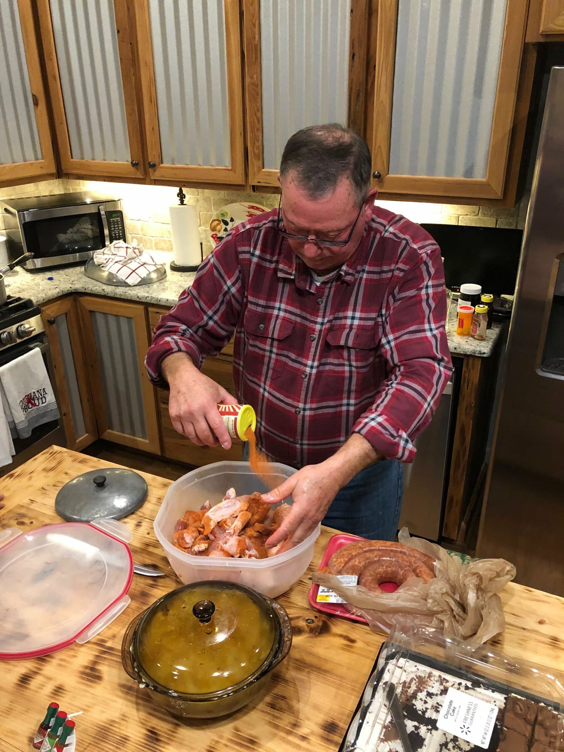 A man in a plaid shirt pours seasoning onto shrimp in a bowl while preparing food in a kitchen.