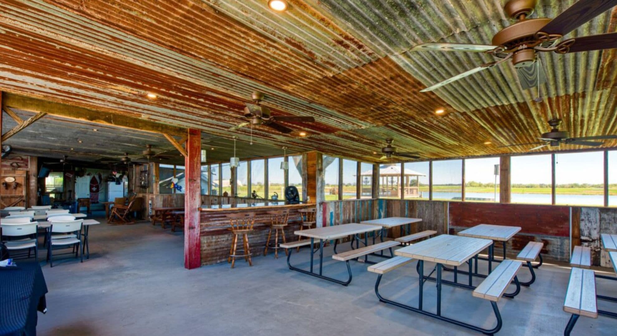 Rustic indoor space with wooden tables, fans, and large windows overlooking a body of water.
