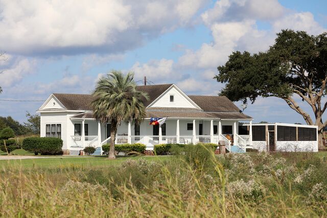 A large white house with a porch, surrounded by greenery and trees, set against a cloudy sky.