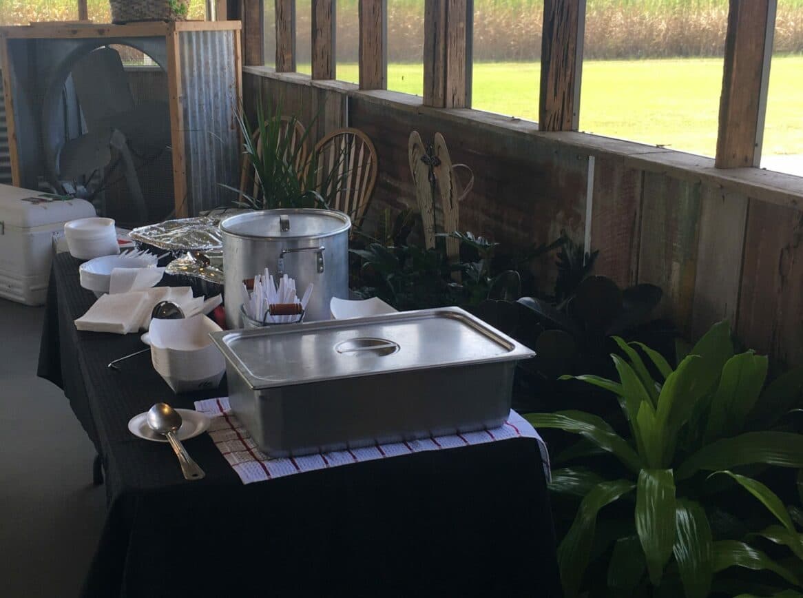 A decorated table with a serving dish, utensils, and disposable containers surrounded by plants and a rustic setting.