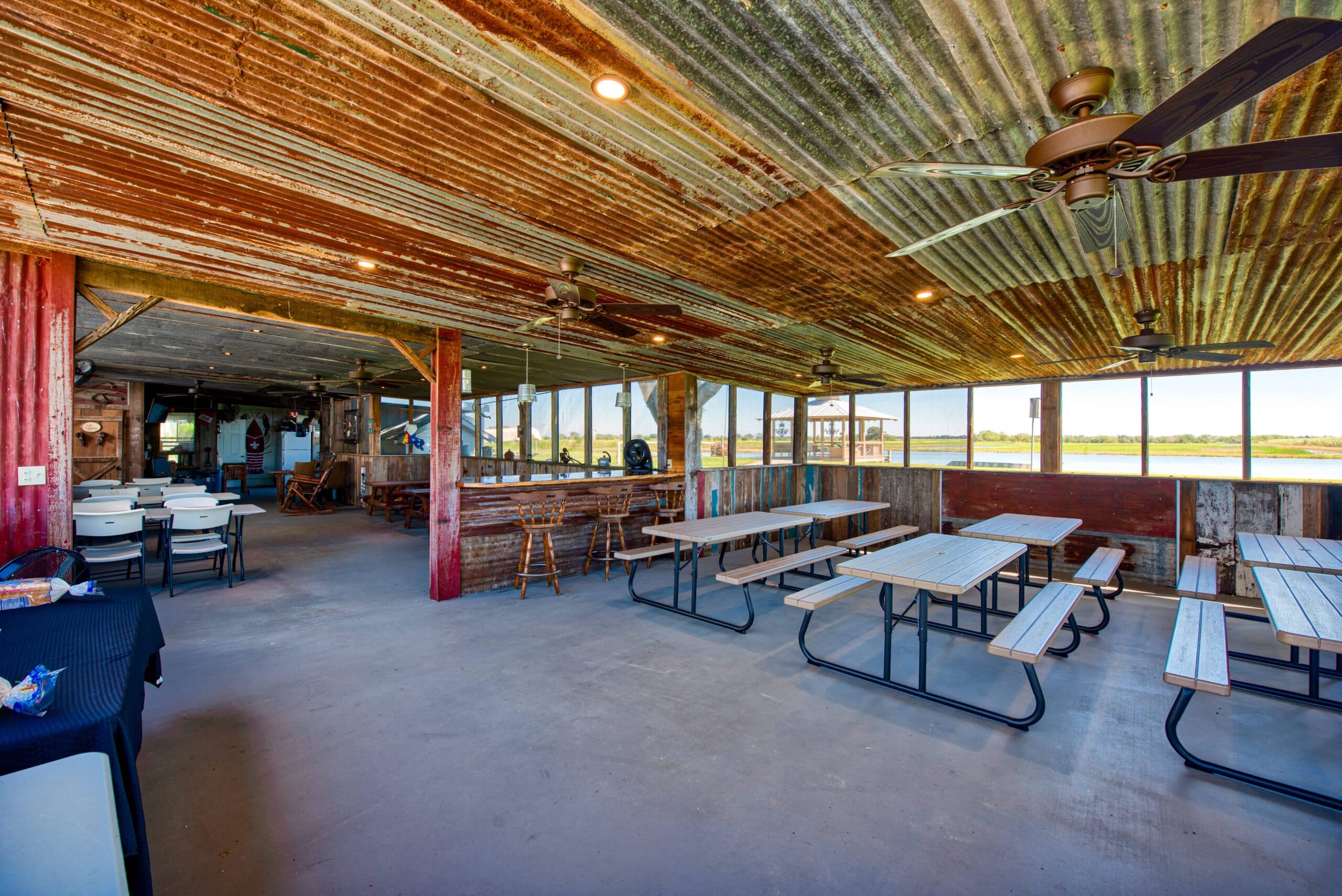 An open, rustic dining area with metal roofing, picnic-style tables, and large windows overlooking a field.