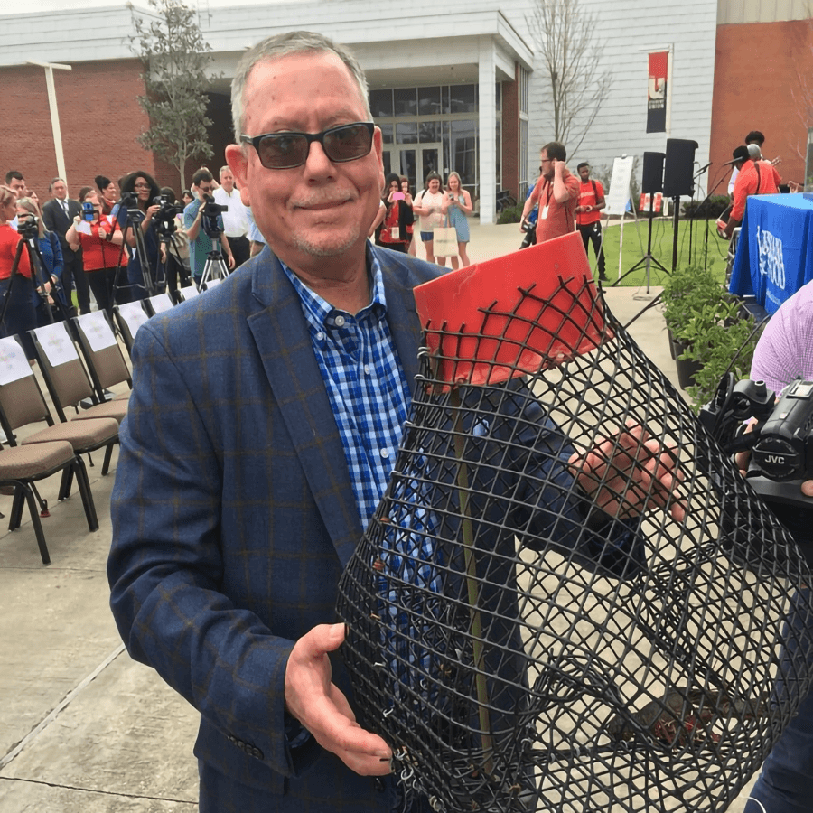 A man in glasses holds a large fish trap at an outdoor event with a crowd in the background.