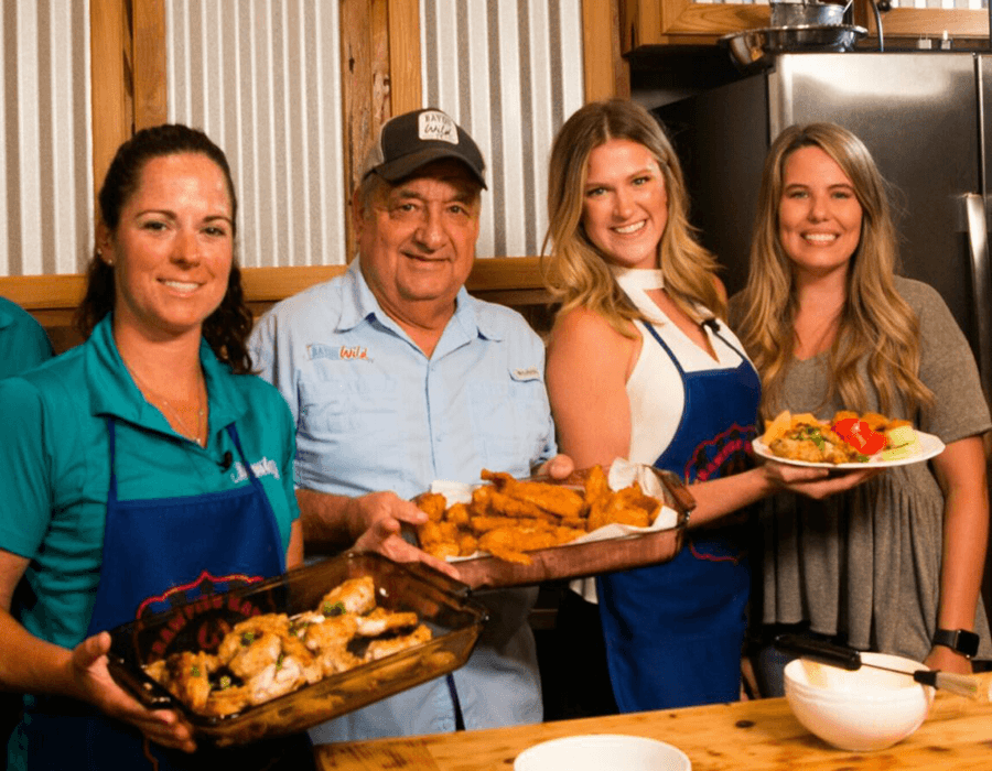A group of four people smiles while holding various plates of food in a kitchen setting.